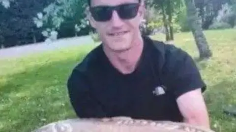 Family Louis Price is wearing a black T-shirt and dark sunglasses. He is holding a large fish close to the camera and smiling. There is greenery and trees in the background.