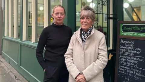 Jonny Humphries/BBC Carly Lea, who has blonder hair tied back and a black jumper and black trousers, stands next to Susan Lea who has blond hair cut into a fringe and a cream trenchcoat. They stand next a chalk board reading 'Maggie May's of Liverpool' followed by a menu. 