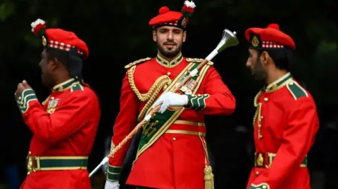 Getty Images Three members The Royal Army of Oman Pipes and Drums practise., They are wearing red jackets and glengarry's. The man in the centre is holding a drum majors baton.