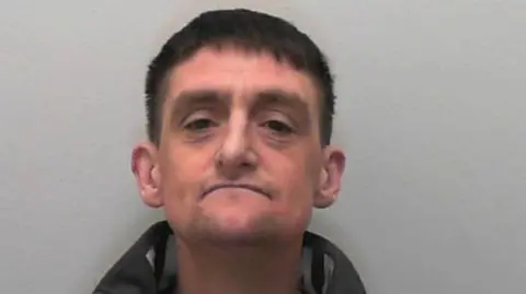 A mugshot of Christopher Bullivant - a man in his 40s with jet black hair and an anorak. He is staring into the camera.