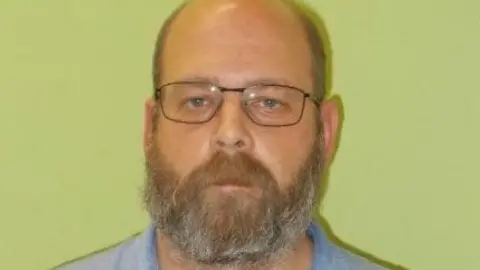 Guernsey Police A mugshot of Benjamin Solowan. He is bald, is wearing glasses, and has a medium length brown and grey beard. He is staring into the camera blankly.