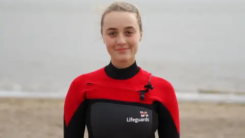 Lois Kemp stood on a beach, smiling at the camera. She is wearing a black and red swimsuit with the RNLI Lifeguards logo on her chest. She has blonde hair tied back. 