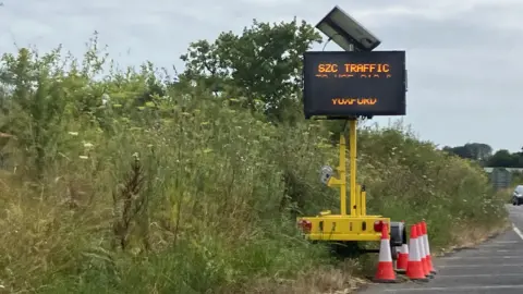 GUY CAMPBELL/BBC A yellow electronic road sign beside the A 12 has messages for "SZC Traffic" 