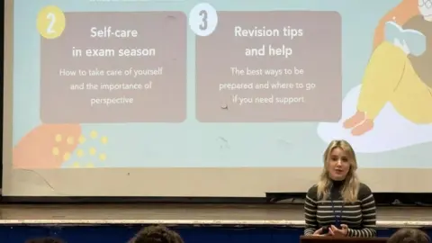 Be Free Campaign A woman with long blonde hair stands in front of a presentation explaining how to manage stress during exam season. She wears a blue and white striped top .