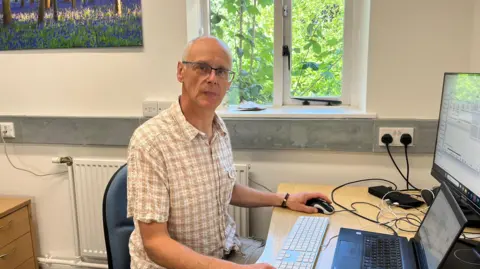 A person sits at a desk in a modern office environment, working on a computer setup that includes a laptop and an additional monitor. Behind them, a window reveals a view of lush greenery, bringing a touch of nature into the workspace. A framed picture of a forest scene with blue flowers hangs on the adjacent wall. The desk is equipped with various electronic devices and cables.
