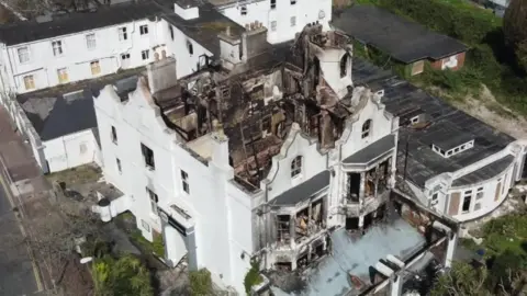 An ariel view of Sherwood Hotel with the burnt roof. The building is white and the inside is fire-damaged. There are white buildings surrounding the area. 