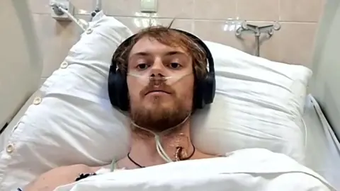 Edward lies in a hospital bed wearing black headphones. He has a tube from his nose and in his neck. His shoulders are uncovered and his head is resting on a white pillow.