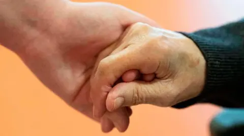 THOMAS KIENZLE/AFP via Getty Images Two people holding hands - one younger hand, one clearly older