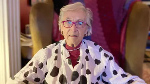 BBC Glenys Cour sitting in a mustard armchair with a pink, red and purple blanket behind her. She has white short hair and is wearing pink glasses, pink earrings and a white blouse with black irregular spots. 