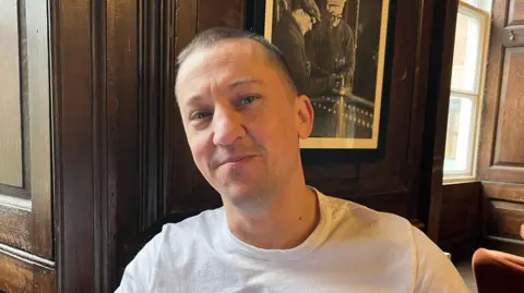 Stefan is looking into the camera and smiling slightly; he is wearing a white t-shirt. He's pictured in a bar lounge area, with some orange armchairs just visible in the right hand corner. Behind him is a dark wood panelled wall, with a black and white photograph on the wall. 