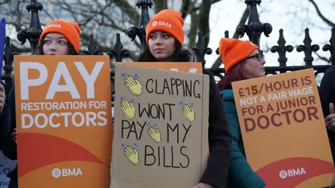 Doctors on strike wearing orange hats with BMA on them, holding placards saying 'pay restoriation for doctors' and 'clapping won't pay my bills'