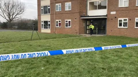 Exterior of a block of flats. Police tape crosses the foreground of the image. A policewoman is standing at the main doors to the flat. A device is standing on a tripod on the path leading out from the doors. The building is surrounded by grass. 