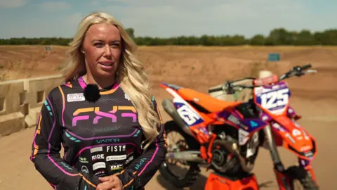 A woman with long blonde hair and wearing a long-sleeved black sports top with white logos stands in front of a large orange motorbike, decorated with large white logos and branding.