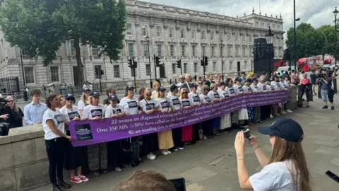 A group of women, all wearing white campaign t-shirts, stand in front of a set of white Portland stone government buildings in Westminster, all holding a long purple and white campaign banner.
