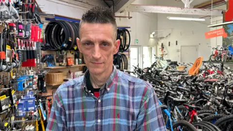 A head-and-shoulders picture of Simon Heard looking directly at the camera. He is wearing a blue checked shirt and has short hair which is shaved at the sides and gelled in small spikes on top. He is standing in front of lots of bikes in a workshop.