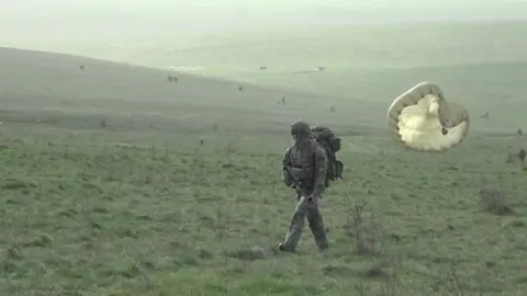 A soldiers walking on the ground after his parachute landing. He is holding a firearm. Others can be seen in the distance behind him.