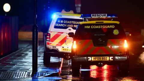 The rear of two HM Coastguard rescue vehicles with flashing lights parked on the street. It is dark and the streets are wet.