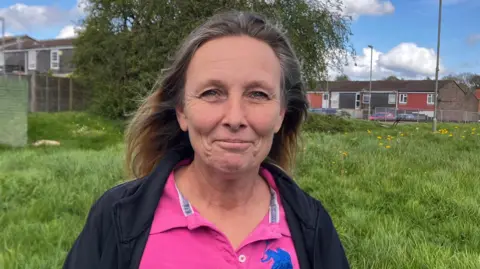 Kelly Inch, Lou's mum near her home in Waterlooville. She has long blonde hair and is smiling. She is standing on a grass area with trees and houses behind. She is wearing a pink polo top and a black cardigan and is smiling at the camera.