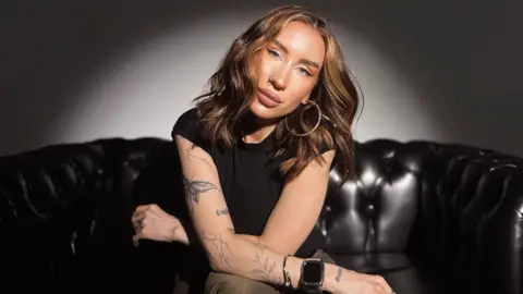 Katya has long mid-brown hair and blue eyes. She is wearing big hoop earrings and has her nose pierced. She has tattooes in the shape of plants and butterflies down one arm. 