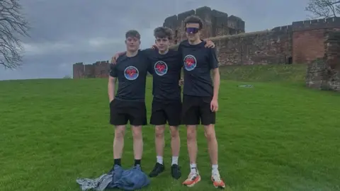 Supplied Theo, Oliver and Jacob standing outside Carlisle Castle with their arms around each other. It is a grey day and there are standing on green grass. They are all wearing black shorts and dark blue tops with a circle symbol on them with a red heart inside. They all have dark brown hair. Jacob is wearing sunglasses.
