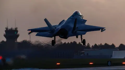 Getty Images A fighter jet takes off from RAF Lakenheath. The light is low and heat coming from the jet can be seen rippling in the area. Other aviation buildings on the site can be seen in the distance. 