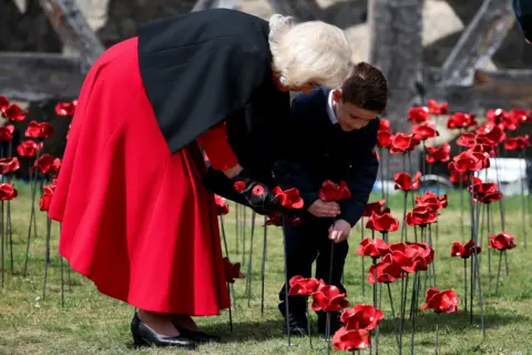 Reuters Queen Camilla bends over to view an installation of ceramic poppies alongside a young boy who is pushing one into the ground