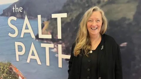 BBC A picture of Raynor Winn stood in front of a banner with the words 'the Salt Path' on it. It is a picture of the coast in the background.