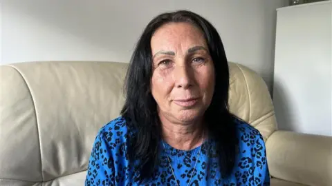 Lisa Bones sits on a cream leather settee in her temporary accommodation. She has black shoulder length hair and is wearing a blue and black leopard print dress, with a silver necklace 