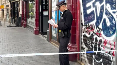 A police officer stands behind police tape on a city street in central London, reading notes outside a closed shop with graffiti on its shutter, while nearby streets are quiet and cordoned off.