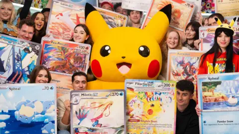 PA Media A large, soft Pikachu - a cut yellow character with rosey red cheeks - is surrounded by young adults holding oversized Pokémon cards.