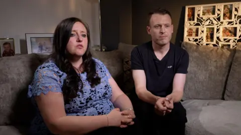 Parents Fiona and Nathan are sat on a grey sofa in their home.