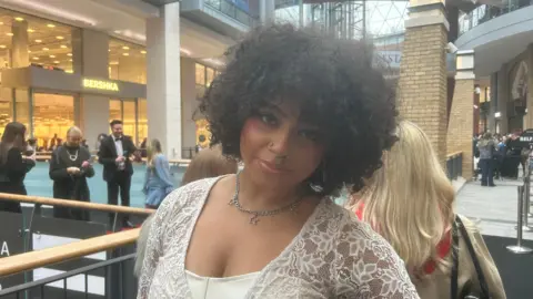 Jade has brown curly hair and is wearing a white long sleeved top. She has a silver necklace and earrings.