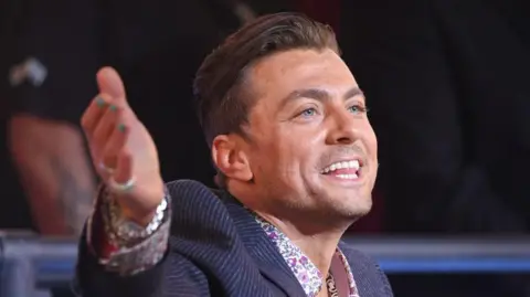 Getty Images Paul Danan wearing a blue pinstripe suit over a pink flowery shirt, a silver watch and several rings, with his nails painted a teal colour. He is smiling at something to the right of the camera and holding out his right hand.
