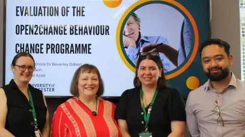 Society Without Abuse Four people - three women, one man - stand in front of a projector screen facing the camera which has a slide displayed saying 'evaluation of the open to change behaviour change programme'