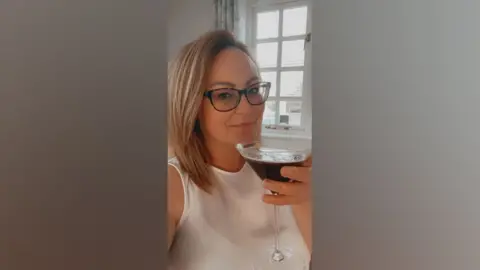 Michelle has shoulder length dark blonde hair, worn with a side parting. She is wearing dark framed glasses and a white sleeveless top and holds a martini-style cocktail glass containing a dark coloured drink.