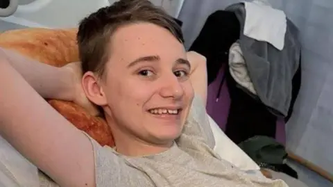 A boy with short brown hair and a grey t-shirt with his arms behind his head