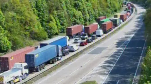 A blurry traffic camera image of the A14. On the right the traffic is stationary or crawling, bumper to bumper with lots of large lorries as well as cars. The right hadn dual carriageway is completely empty. 