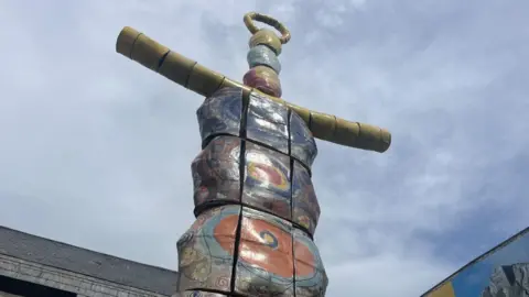 The image shows the top of a very tall statue called the Earth Goddess. It resembles a brightly coloured totem pole and has yellow arms. The sky behind it is blue with thin white cloud. The tops of two buildings are visible too.