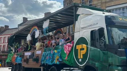 Carroll Weston/BBC People wearing colourful outfits stand inside an open trailer on the back of a lorry. There are rainbows and big banners on display as they pass through the town centre.