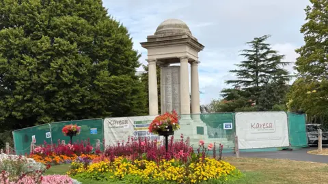 A large stone war memorial with columns and a domed roof in a park. There are large evergreen trees around it and a vibrant flower bed in front. The monument is surrounded by green fencing put in place for the works. 