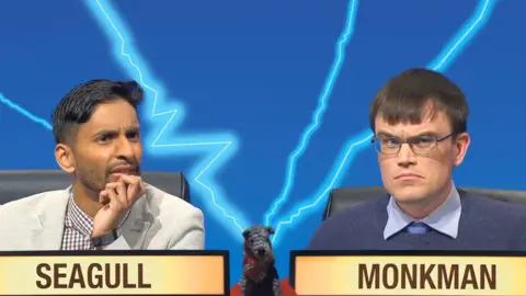 Bobby Seagull, a man with short black hair, wearing a checked shirt and an off-white blazer, sits next to Eric Monkman, a man with short brown hair, wearing a glasses, a blue shirt and a blue jumper. They are on the set of University Challenge, and each has a sign with his name on in front of him.