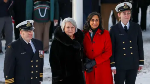 Reuters An image showing Foreign Minister Anita Anand and Governor General Mary Simon, with Canadian officers at either of their sides. Anand is wearing a bright red coat and black gloves, and she has shoulder-length wavy black hair. Simon is wearing a large fur black coat, and has a short white bob. 