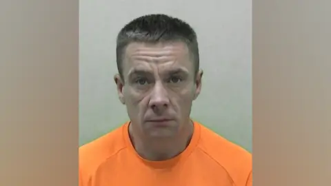 A mug shot of Christopher Donnison. He has short brown hair and is wearing an orange t-shirt. He is staring into the camera with a straight face.