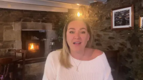 A blonde haired woman wearing a white sweater standing in front of a fire in a pub