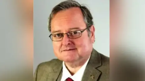 Harris is in a green/brown suit and a white shirt He is looking at the camera and wearing lasses. He has short, brown hair.