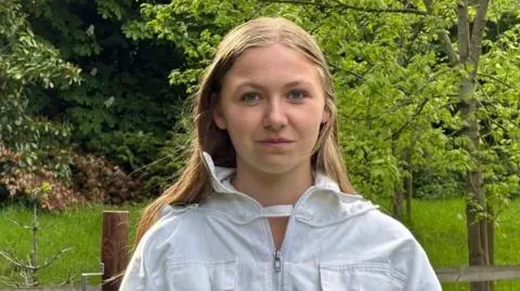 Charlie Stubbs / BBC There is a 16 year old girl who is wearing a white beekeepers suit. She has light hair and brown eyes. She is stood in front of a wooden fence, with a pillar over one of her shoulders. Over the other, there is a thin tree with light green leaves.