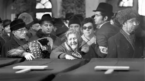 Women mourn the victims in a black and white photo showing coffins