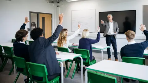 Children sitting in a classroom on green chairs with their hands up. A teacher is standing in front of a whiteboard and large black television screen. He is pointing towards the class. 