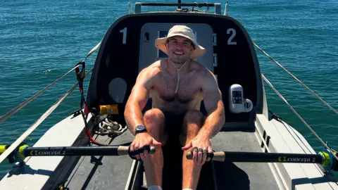Olly Rathbone, in a cream-coloured wide-brimmed boonie hat, rows a boat before attempting to cross the Atlantic 