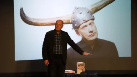 Westlands Adrian Edmondson speaking on stage at the Yeovil Literary Festival, with a large picture behind him on a screen of him wear a hat with large horns